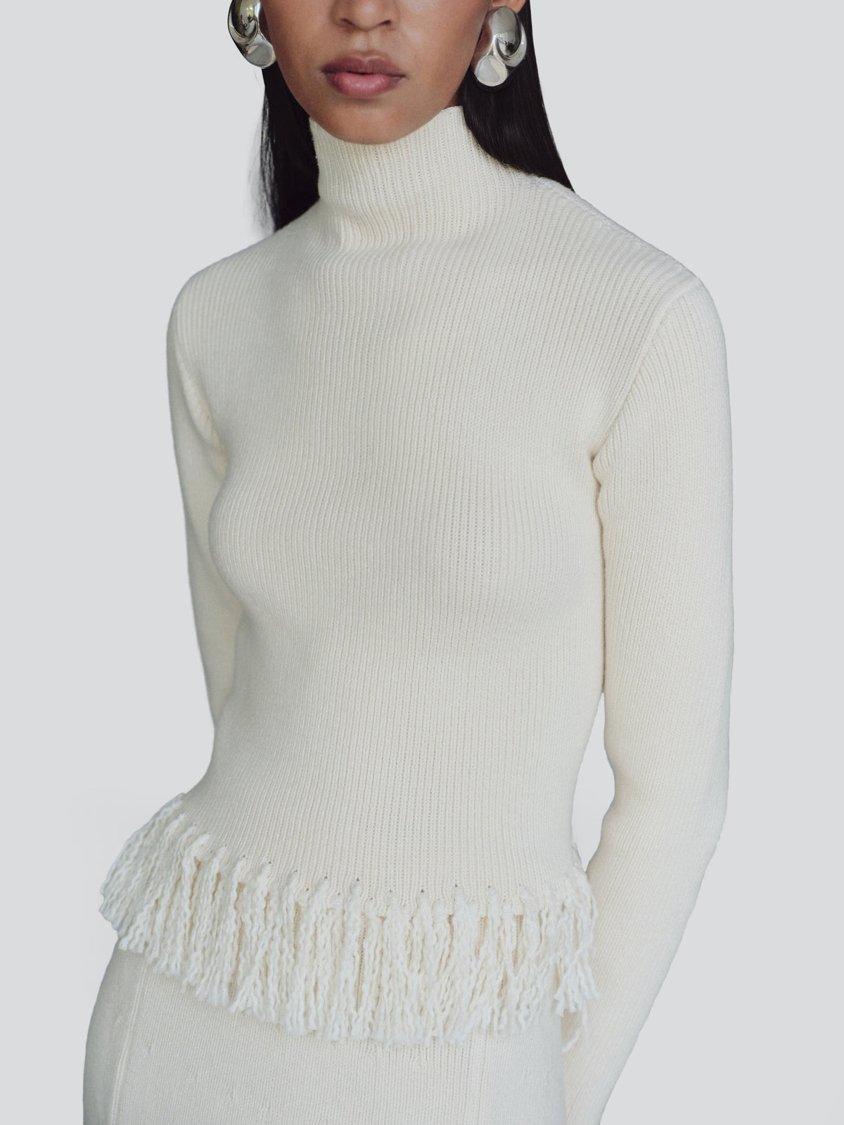 A close-up of a model wearing a ribbed cream turtleneck sweater with a fringed hem, paired with matching cream knit bottoms against a light gray background.