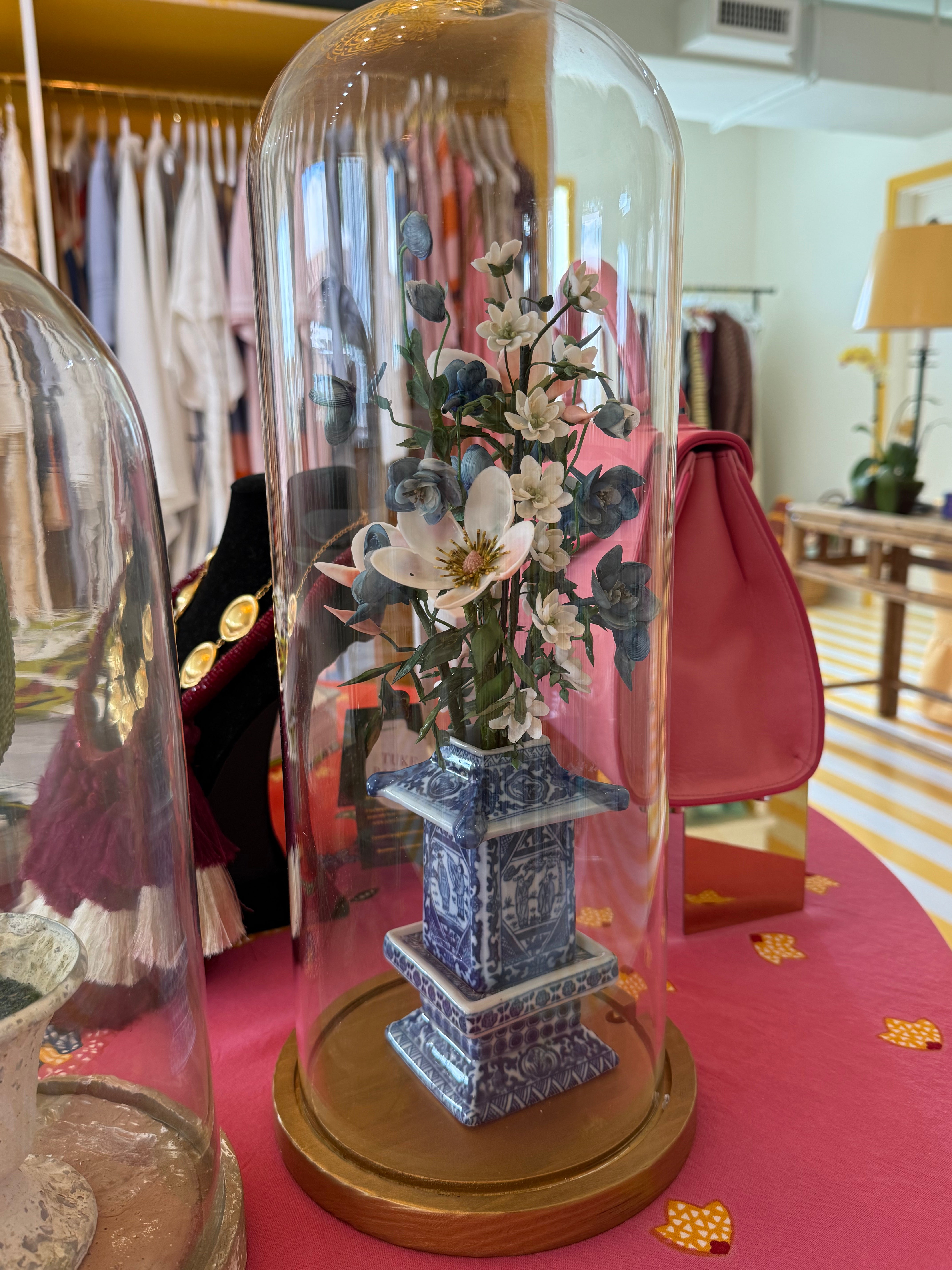 Tall floral arrangement under clear glass cloche with blue-and-white patterned pedestal pot.