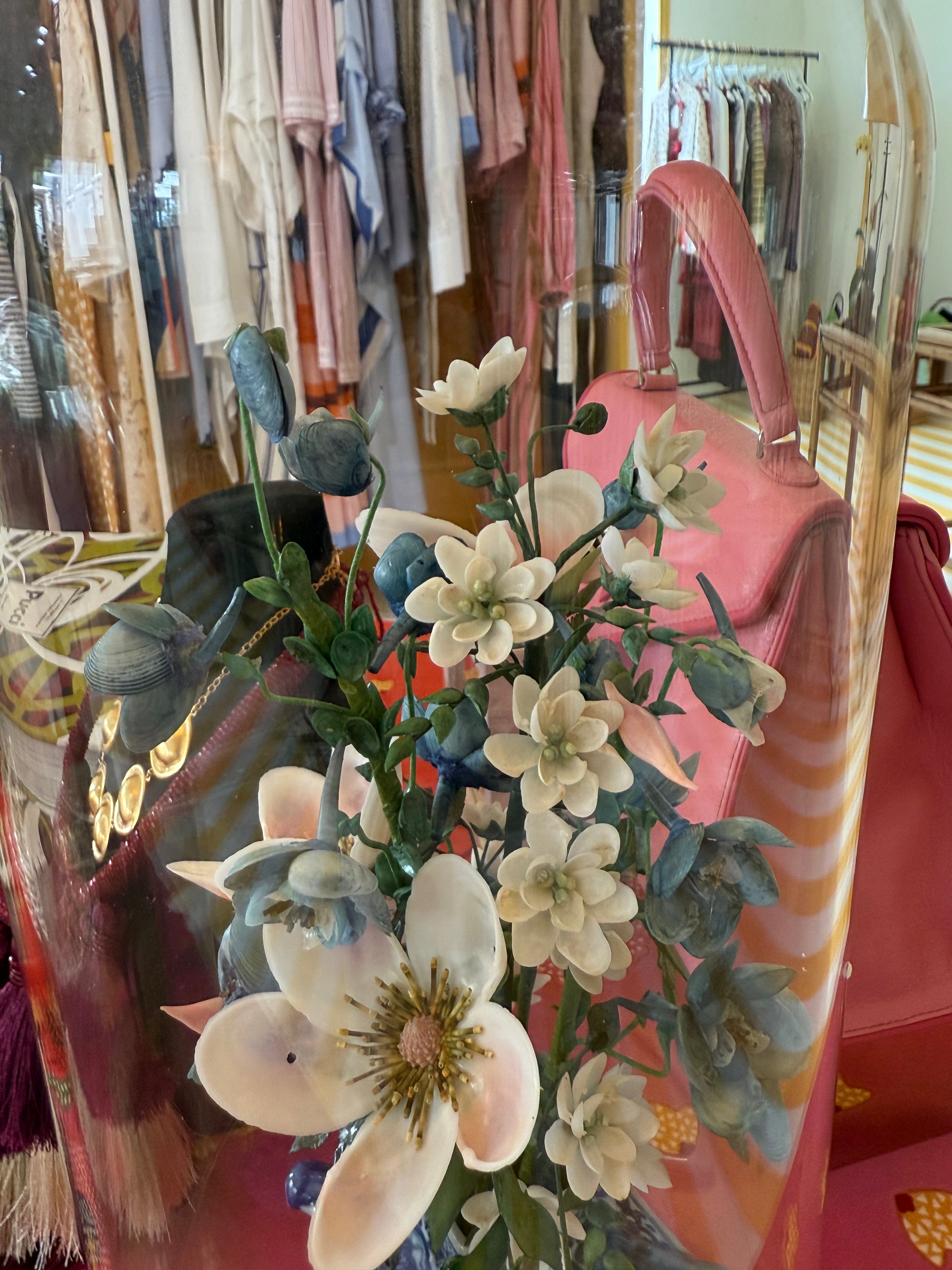 Floral arrangement under clear glass dome with white flowers and blue foliage on a blue pot.
