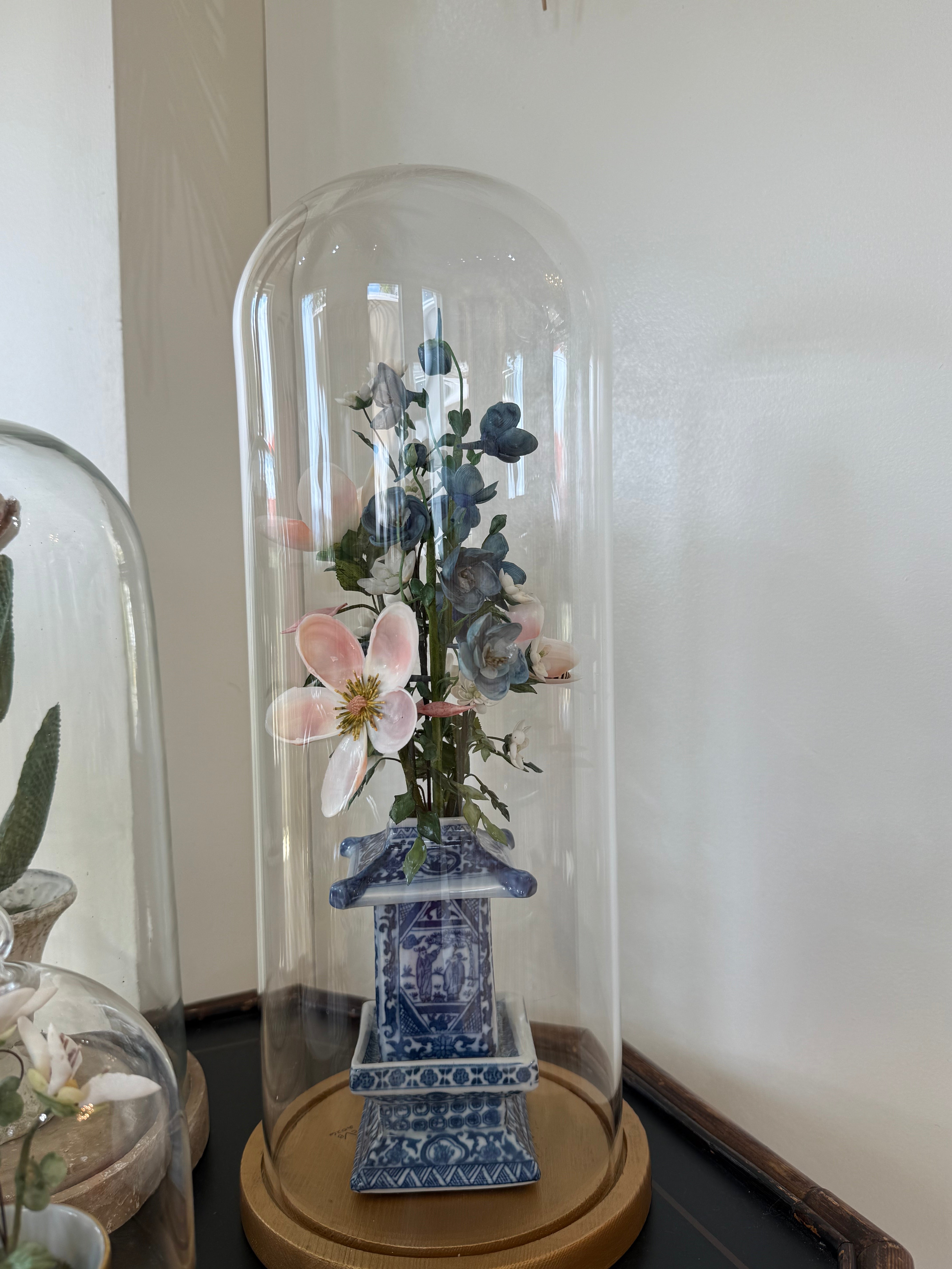 Blue and white floral arrangement under clear glass dome with porcelain pedestal pot.